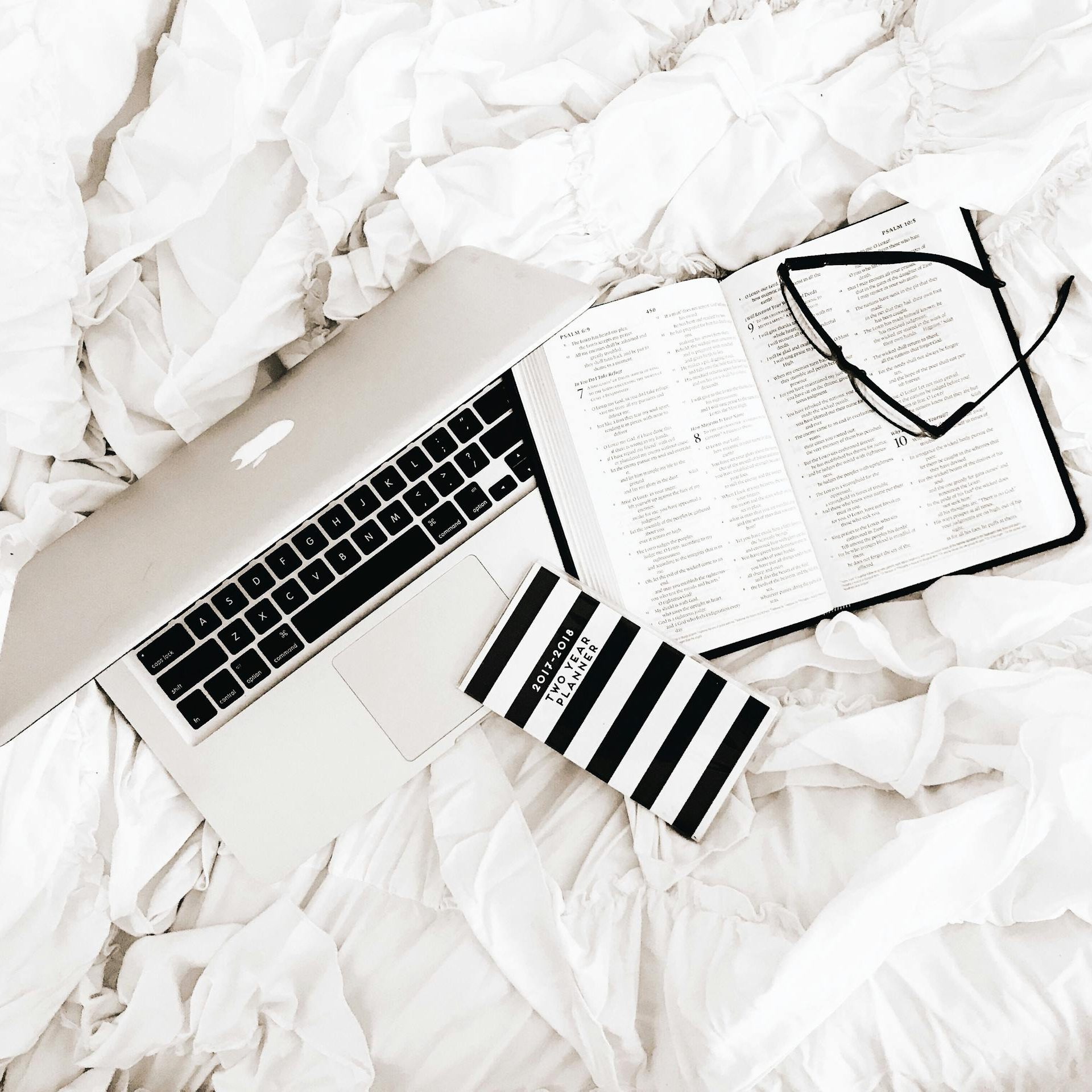 A flat lay of a laptop, open book, and reading glasses on a white bed. Perfect for productivity and relaxation themes.